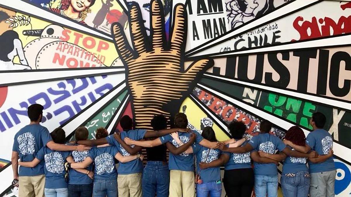 A group of students stand together in front of a mural with arms over each others shoulders