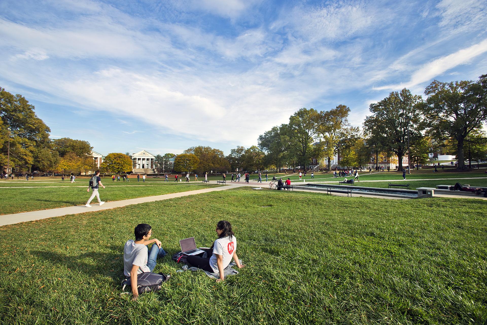 A group of students sit on the green grass of McKedlin Mall under a blue sky